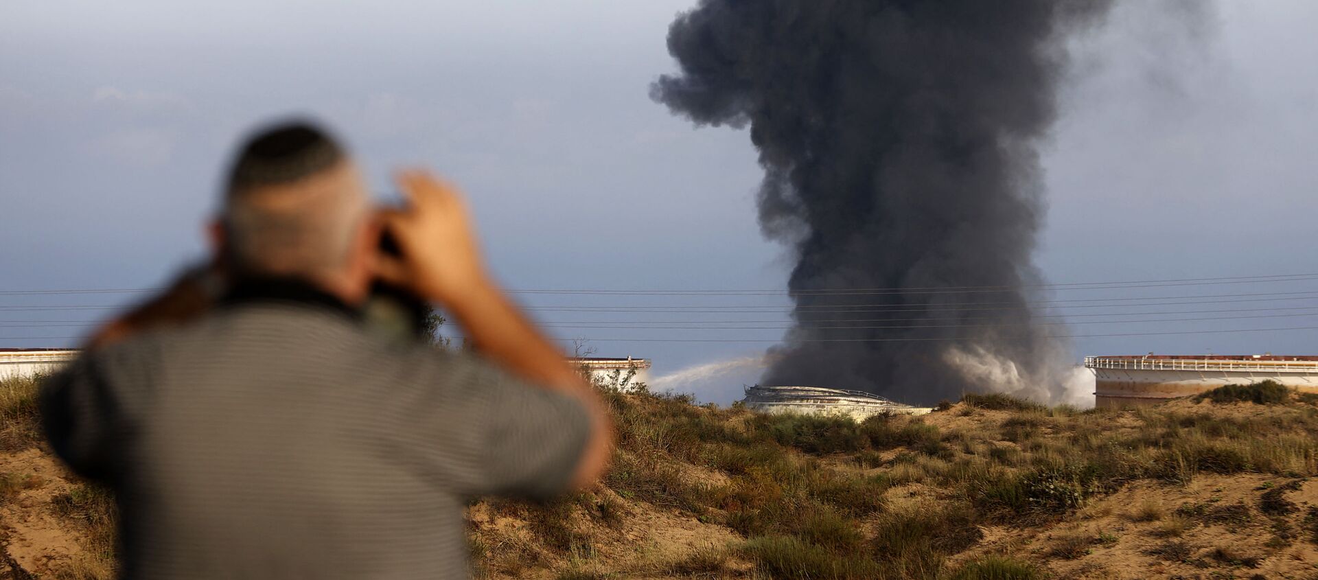 Một người đàn ông Israel nhìn từ xa khi khói bốc lên vào sáng sớm ngày 12 tháng 5 năm 2021 từ nhà máy lọc dầu của Ashkelon, bị trúng tên lửa Hamas vào ngày hôm trước, ở thành phố phía nam Israel, gần Dải Gaza - Sputnik Việt Nam, 1920, 17.05.2021
