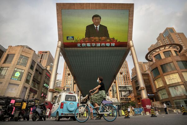 Một người phụ nữ đeo khẩu trang đi xe đạp qua màn hình TV lớn tại trung tâm mua sắm ở Bắc Kinh đang chiếu một phóng sự truyền hình Trung Quốc về chuyến thăm Hồng Kông của Chủ tịch Trung Quốc Tập Cận Bình. - Sputnik Việt Nam