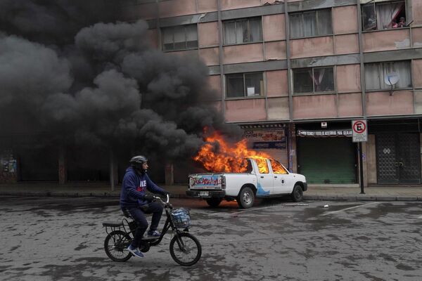 Người đàn ông đi xe đạp ngang qua chiếc xe công vụ của chính quyền thành phố bị đốt cháy ở Santiago, Chile. - Sputnik Việt Nam