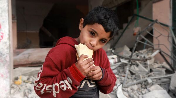 Một đứa trẻ ăn trước một tòa nhà bị hư hại trong vụ đánh bom của Israel ở Rafah - Sputnik Việt Nam