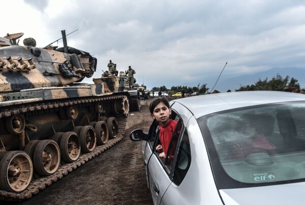 Bé gái Thổ Nhĩ Kỳ ló đầu qua cửa sổ xe hơi nhìn xe tăng và binh lính của quân đội Thổ Nhĩ Kỳ tập trung ở thành phố Hassa, gần biên giới Syria - Sputnik Việt Nam