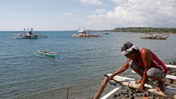 Một ngư dân đang sửa thuyền của mình, trông ra xa là  chiếc thuyền đánh cá đang hoạt động ở bãi biển tranh chấp ở Biển Đông, tại Masinloc, Zambales, Philippines - Sputnik Việt Nam