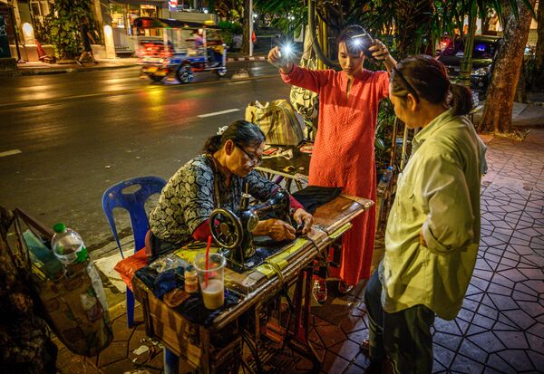 Người phụ nữ bật đèn điện thoại di động tại nơi làm việc của một thợ may đường phố ở Bangkok, Thái Lan - Sputnik Việt Nam